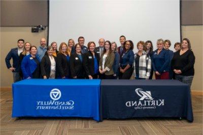 large group of people standing in two rows behind 2 tables draped with banners for GVSU and Kirtland Community College